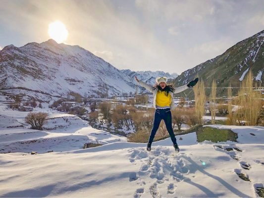 A picture on a woman jumping on snow, sun shining behind, mountain range